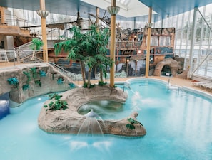 An indoor pool with a waterfall feature, surrounded by artificial rock formations and palm trees. - Holiday Club Turun Caribia (Turku)