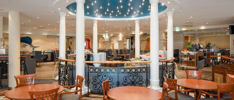A hotel lobby with a central bar area, multiple seating arrangements, and a kitchen in the background.