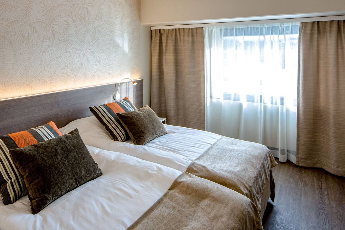 A hotel room with two beds, a headboard, a bedside table, and a window with curtains.