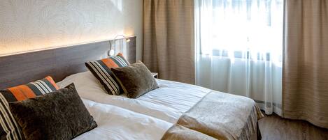 Habitación de hotel con dos camas, cabecero, mesita de noche y ventana con cortinas.