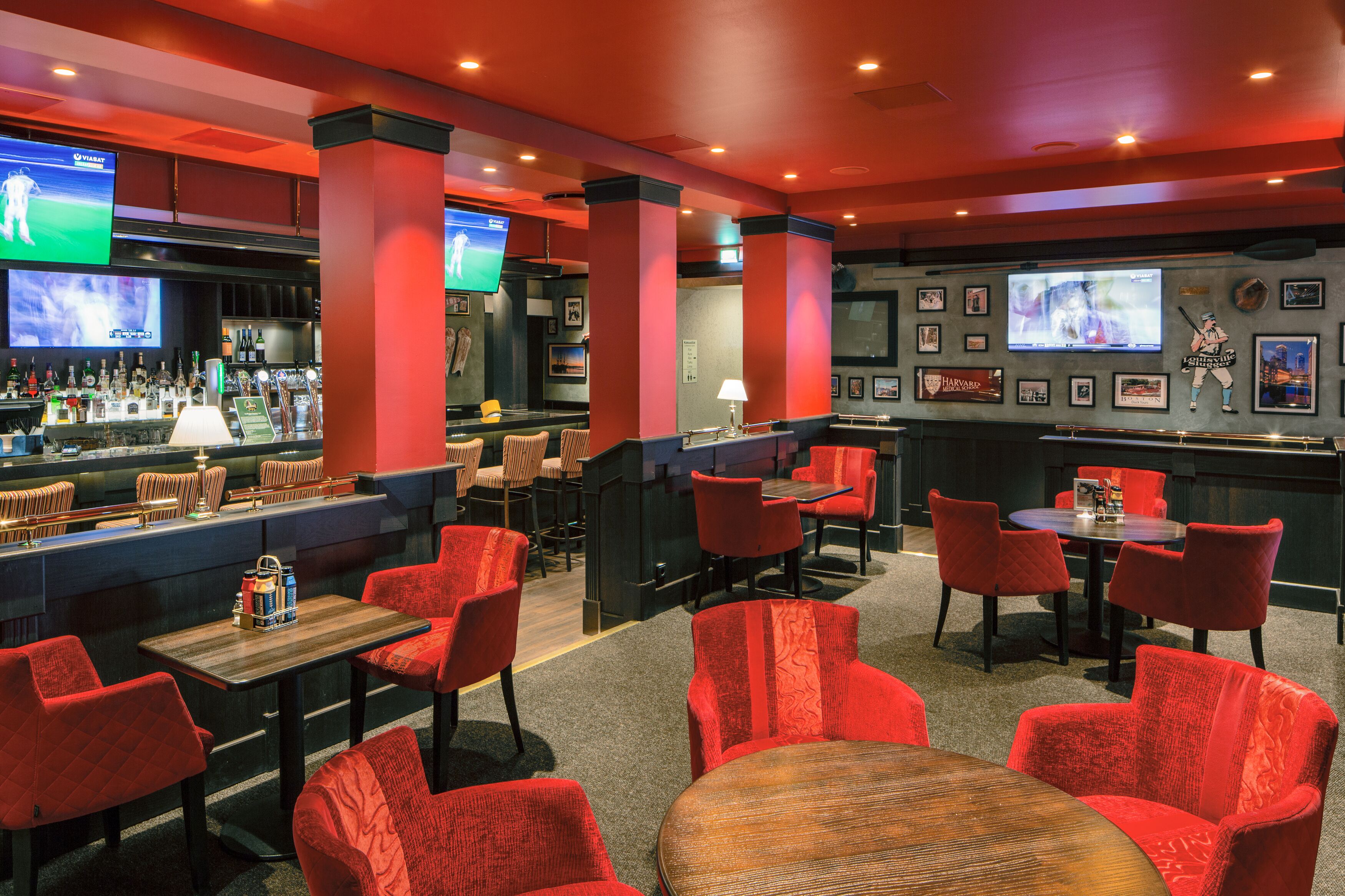 Zona de bar con sillas tapizadas en rojo, una barra con botellas y varias pantallas mostrando eventos deportivos.
