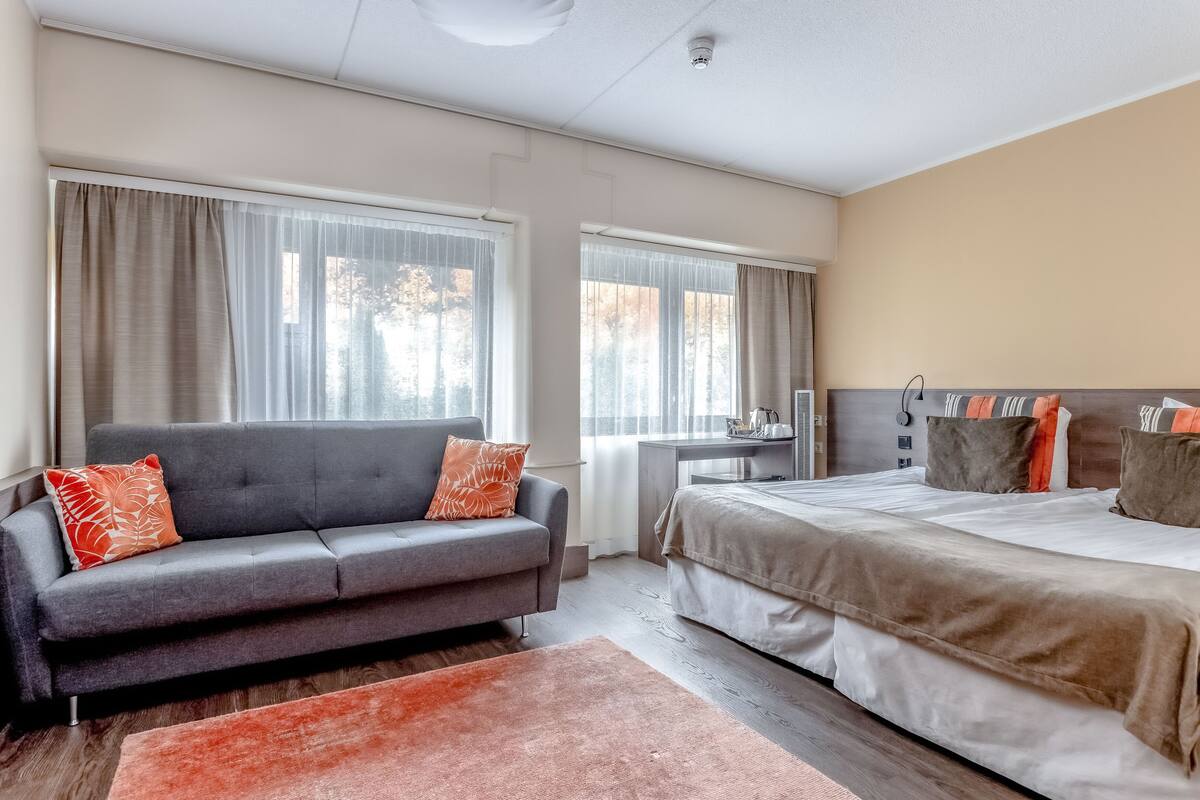 A modern hotel room with a grey sofa, a large bed, and a desk.