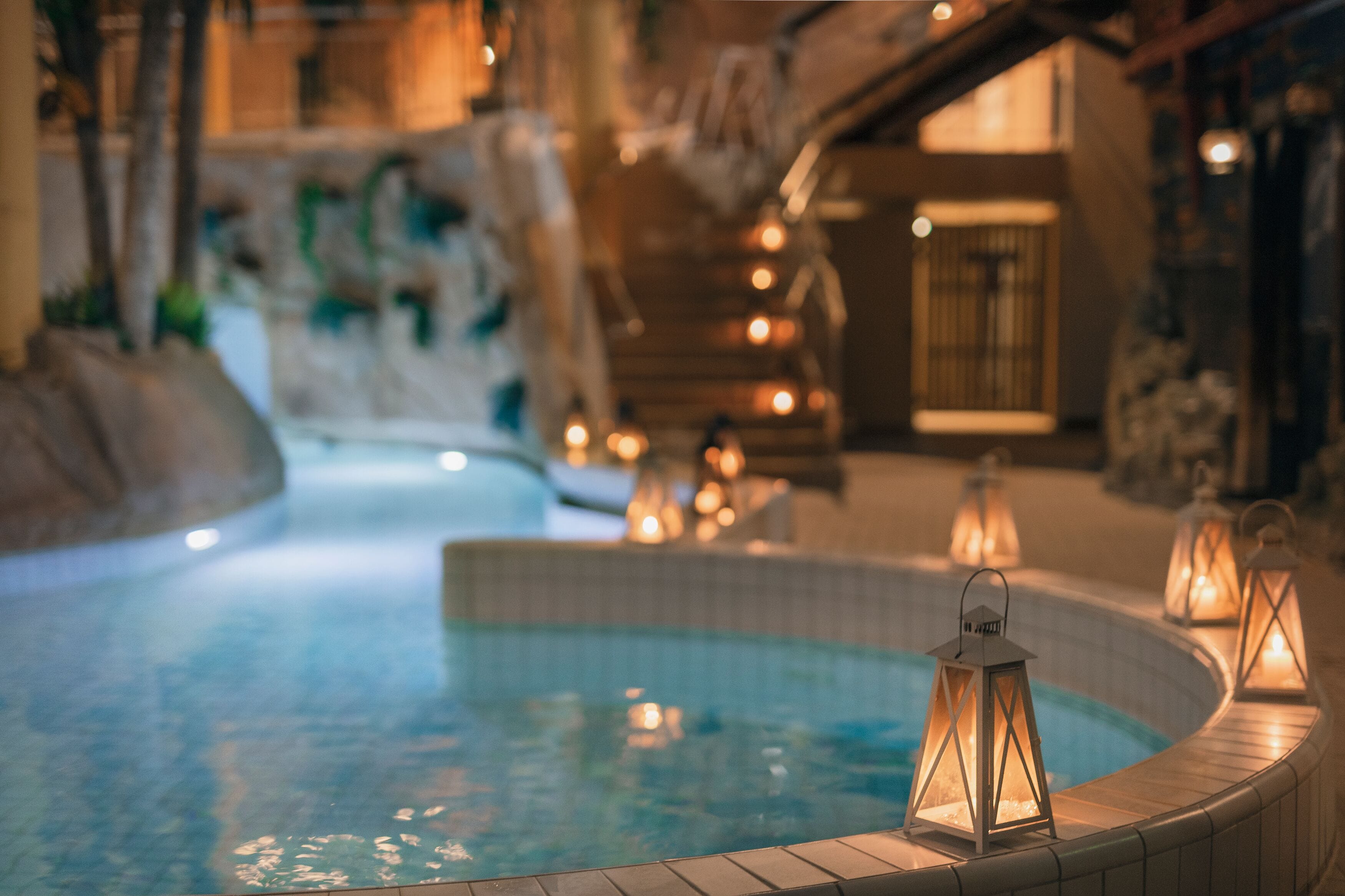 Un área de piscina con borde curvo, iluminada por faroles, y una pared de piedra al fondo.