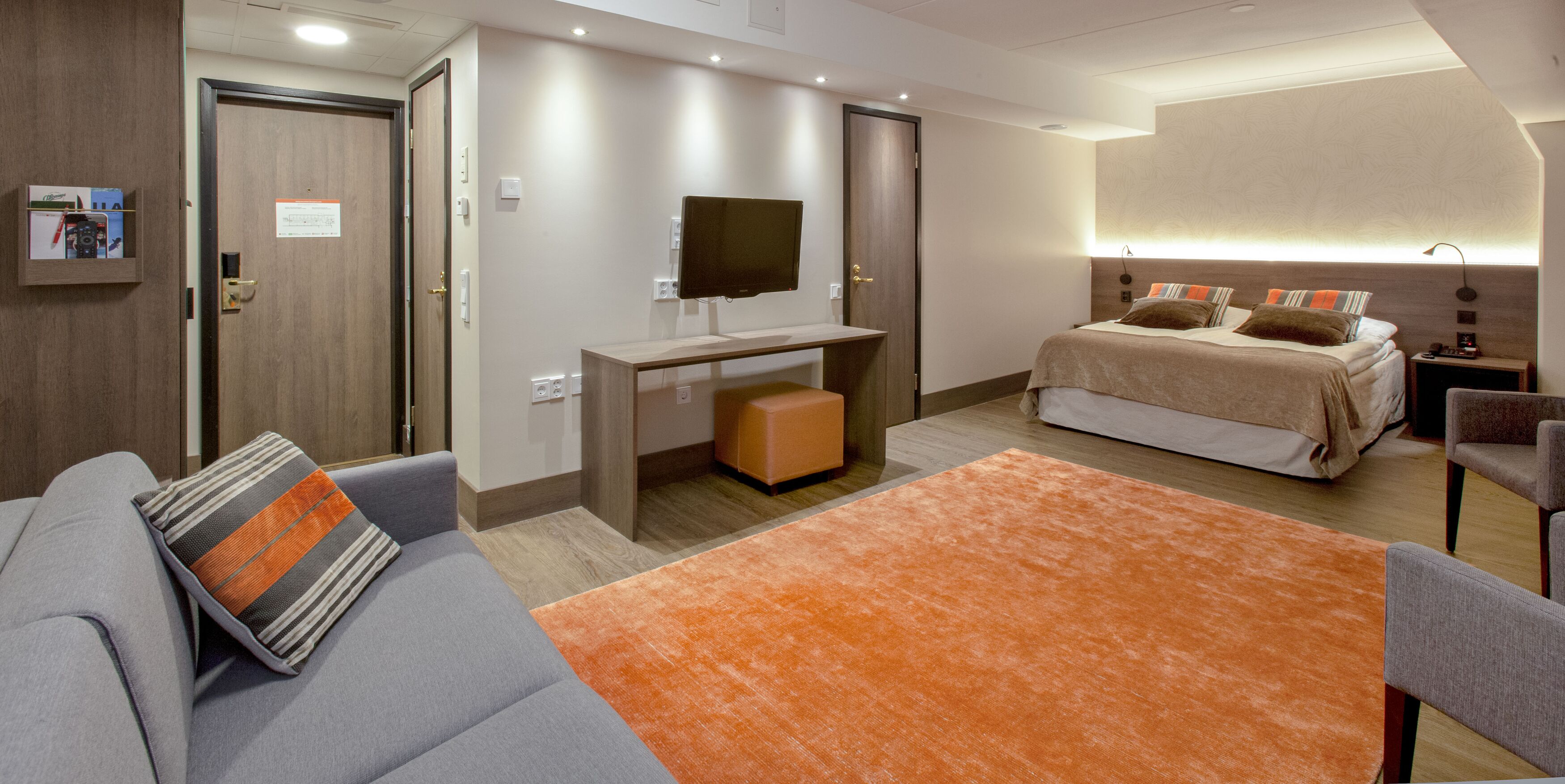 Une chambre d’hôtel moderne avec un canapé gris, un tapis orange, un lit avec plusieurs oreillers, un téléviseur posé sur un meuble en bois et un petit bureau avec une chaise.