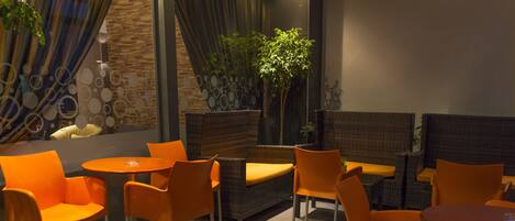 A modern lounge area with wicker furniture and orange chairs, featuring a large potted plant and a stone wall.