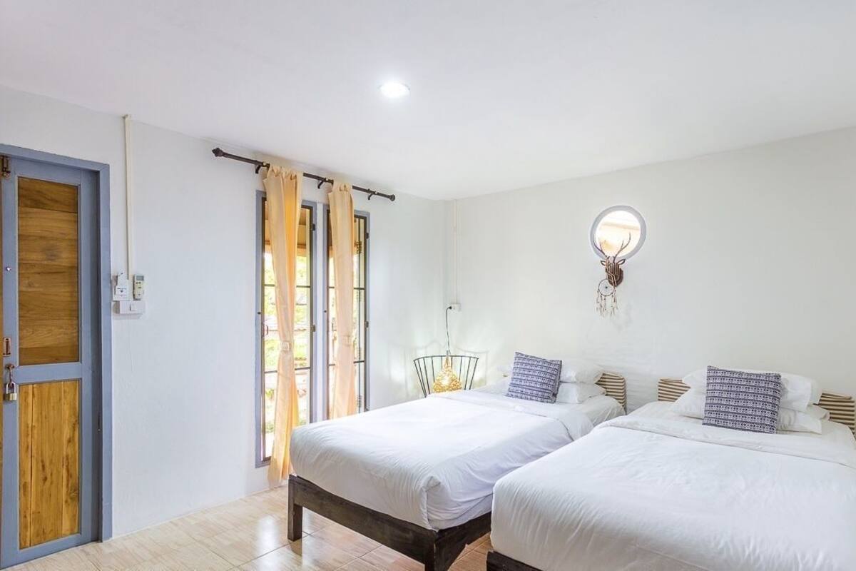 A bedroom with two beds, a window with curtains, a wall-mounted light fixture, and a door.