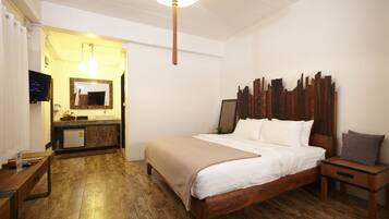 A hotel room with a wooden floor, a bed with white bedding, a nightstand, a bedside table, a mirror, a sink, and a television.