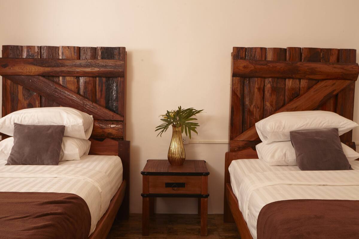 A room with two beds, each with a wooden headboard, a small bedside table, and a potted plant.