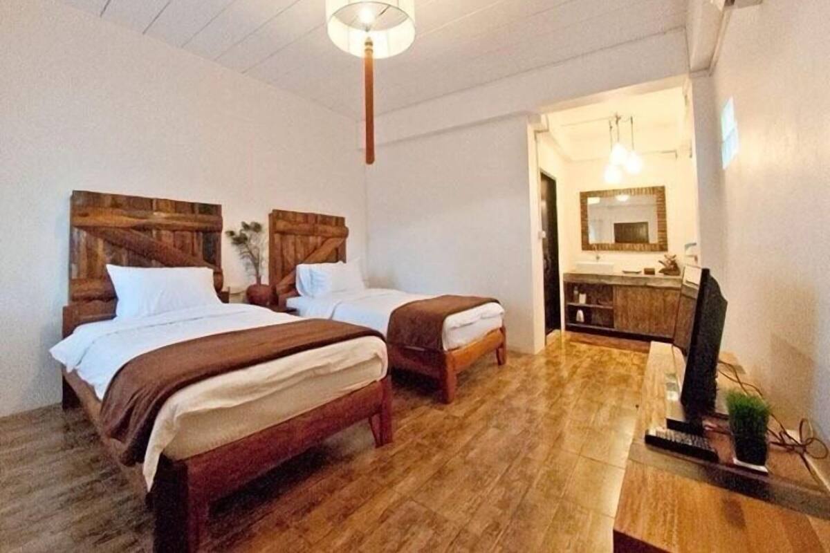 A room with two wooden beds, a wooden table, a television, and a bathroom visible through an open door.