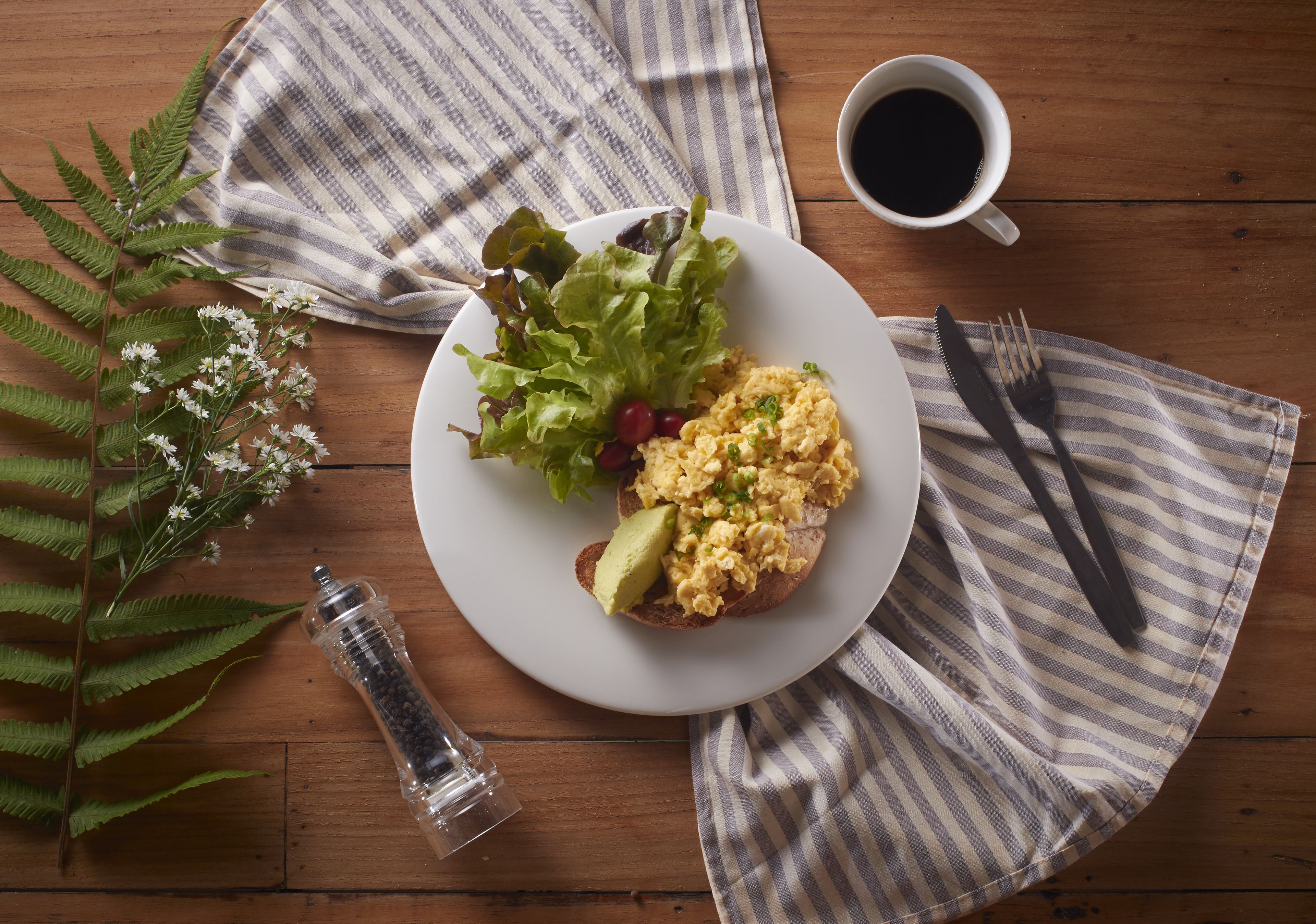 A plate with scrambled eggs, avocado, and lettuce on a wooden table with a striped napkin, a salt shaker, and a cup of coffee.