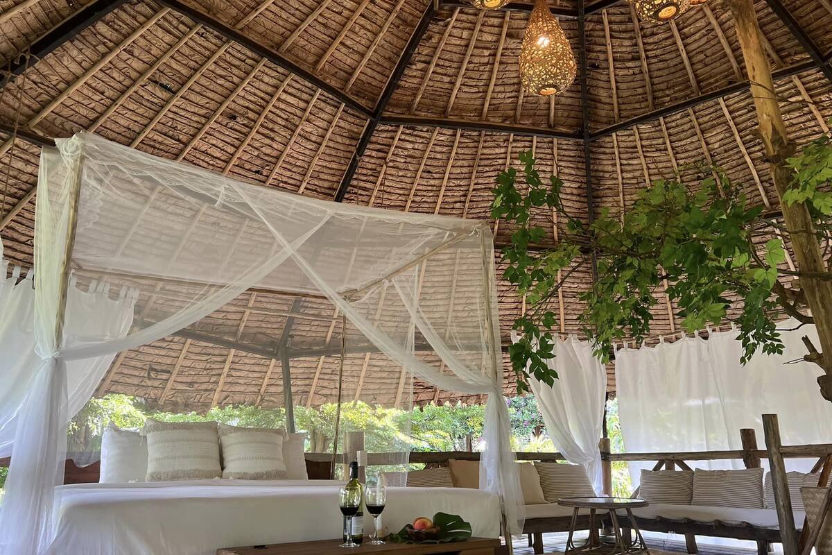 A thatched-roof tent with a canopy bed, a wooden table with drinks, and a seating area.