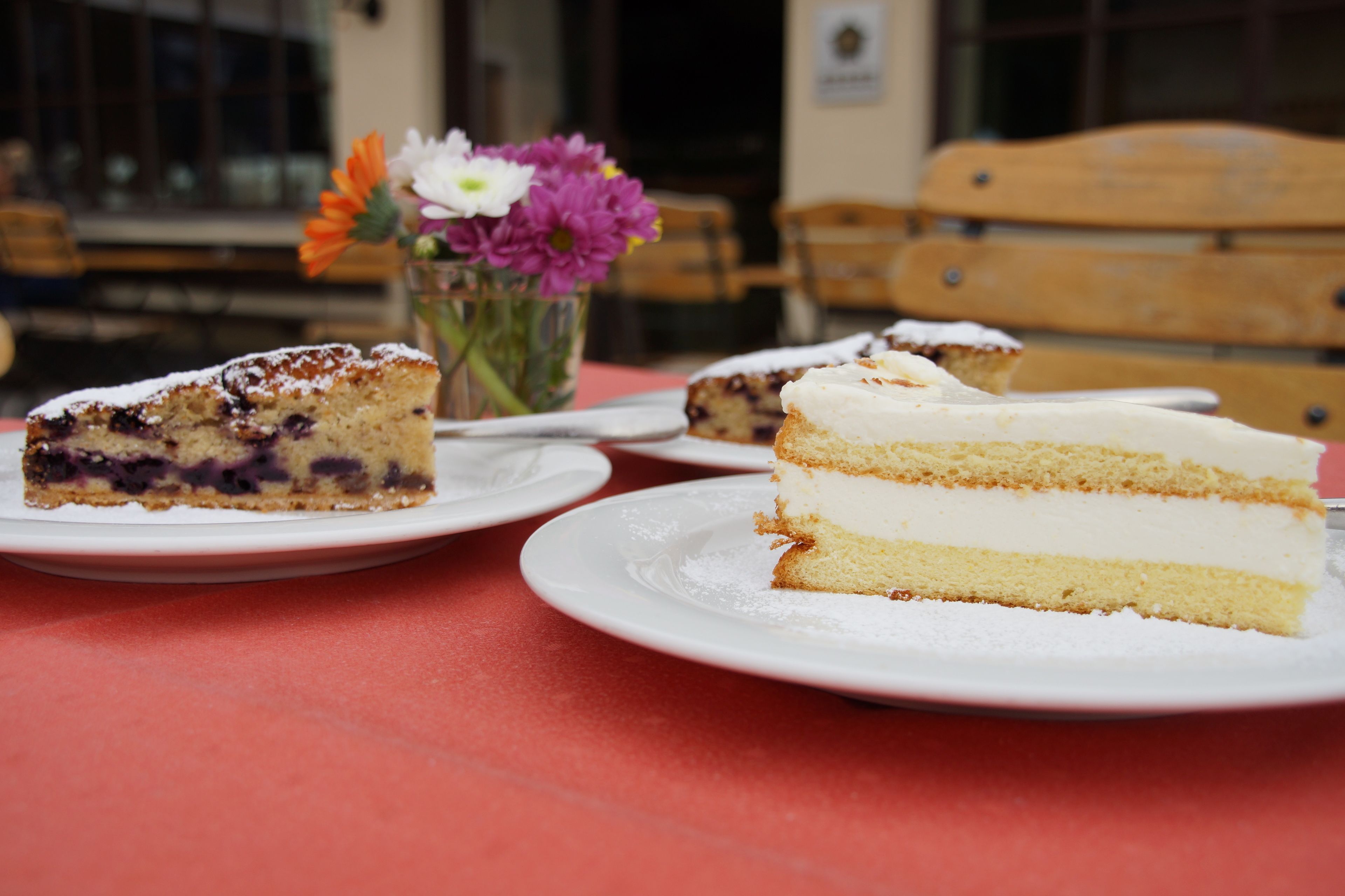 A table with three white plates, each holding a slice of cake. There is a vase with flowers in the background.