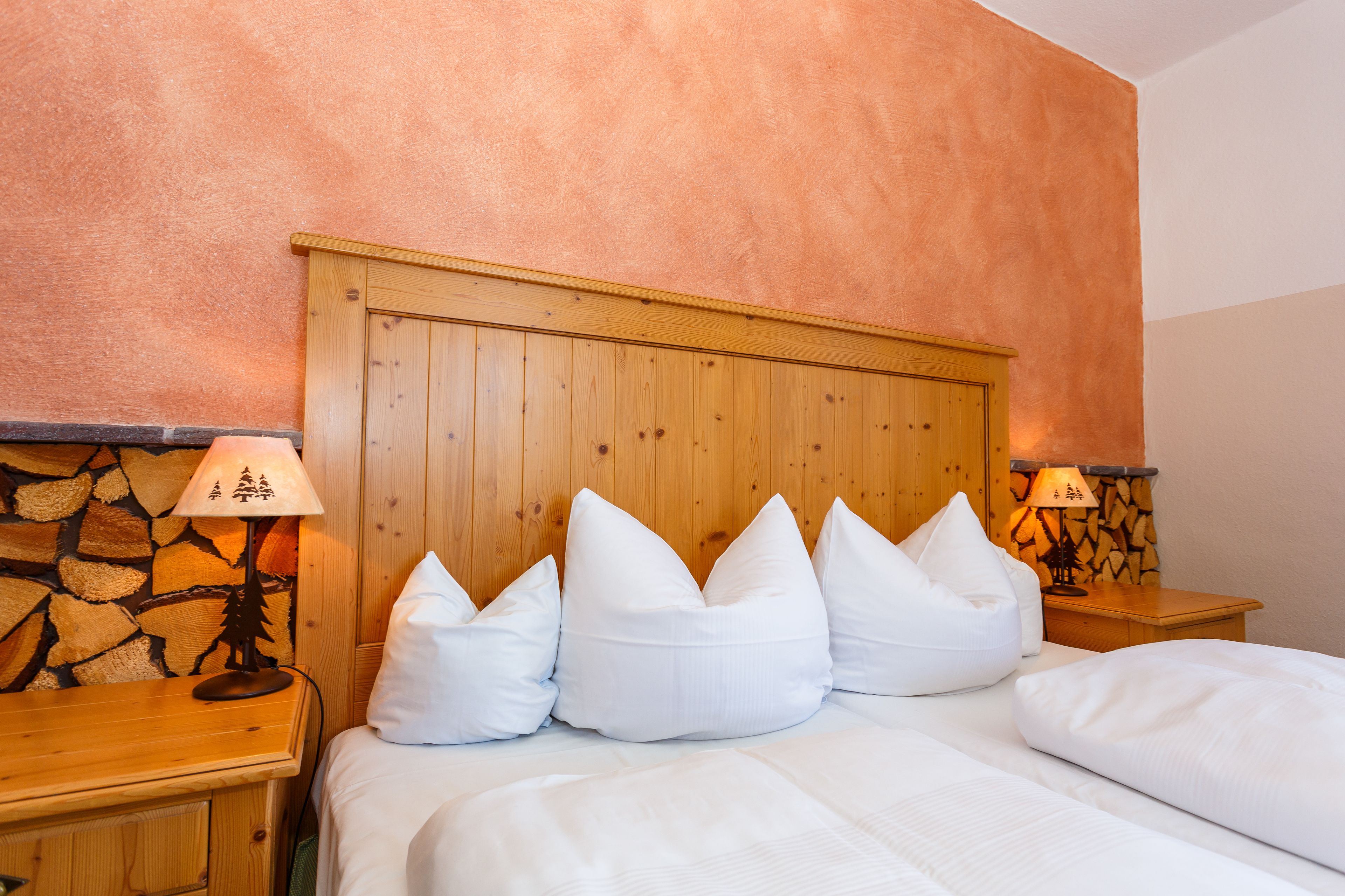 Una cama bien tendida con ropa blanca y varias almohadas, cabecera de madera y dos lámparas de noche con motivos de árboles.