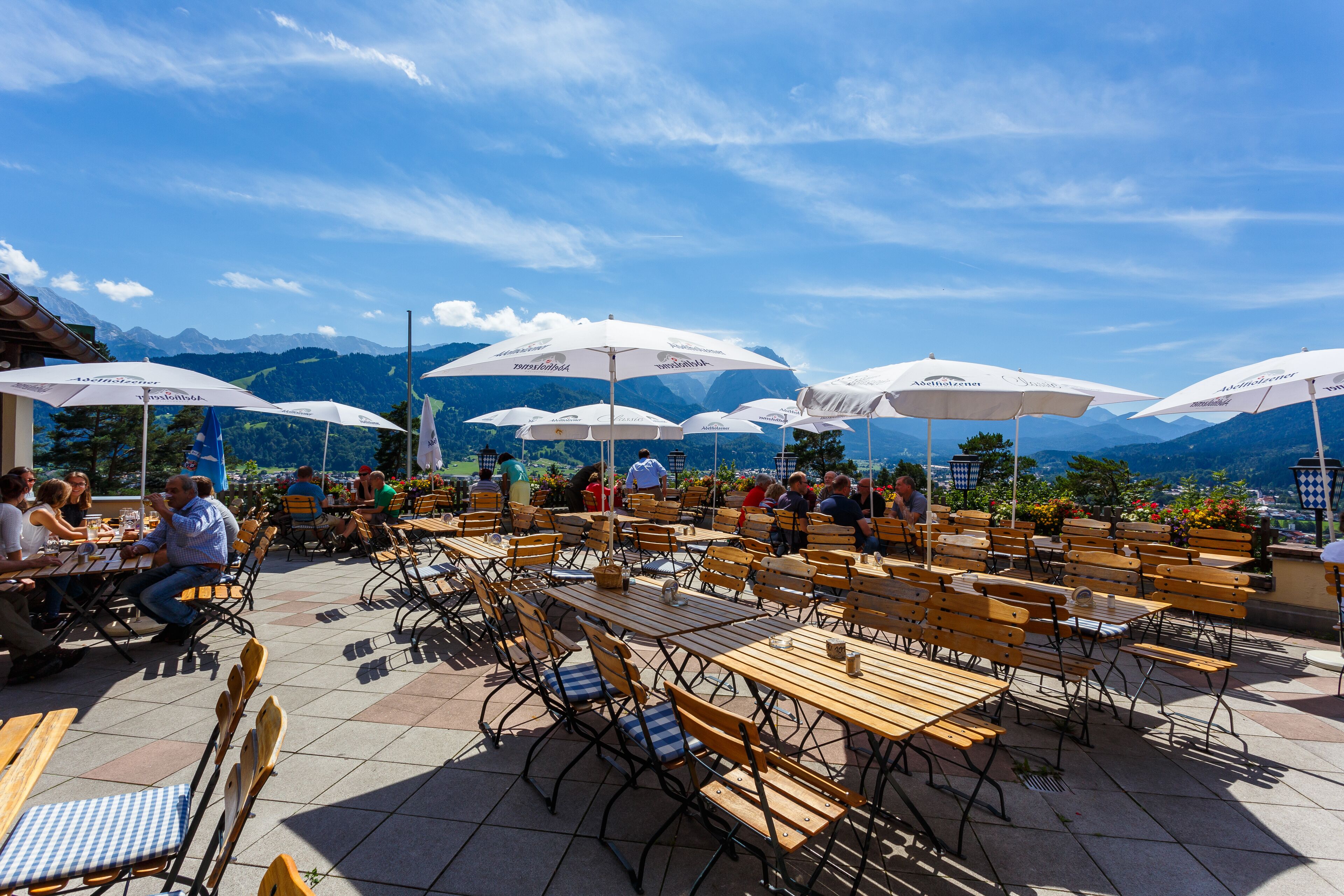 An outdoor café with wooden tables and chairs, white umbrellas, and a mountainous backdrop.