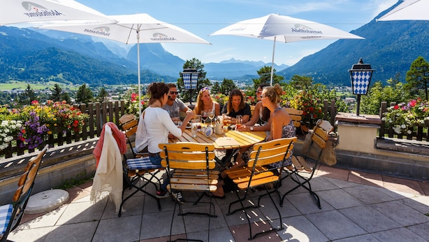 Un groupe de personnes est assis à une table en plein air, profitant de la vue sur les montagnes, à l’ombre de parasols blancs, et une clôture en bois délimite l’espace.