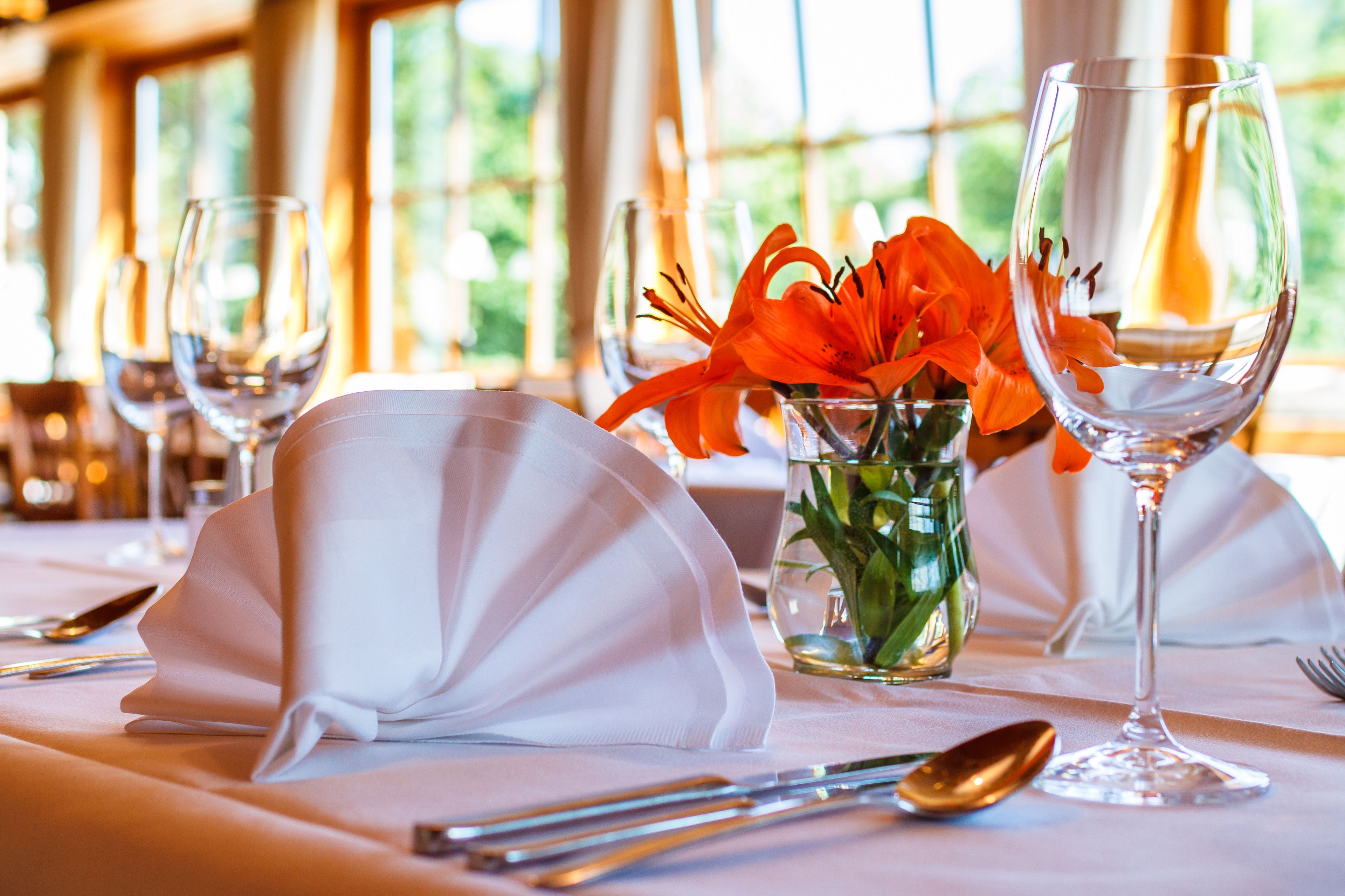Une table dressée pour le repas, avec des serviettes blanches, un vase en verre contenant des fleurs oranges et des verres à vin.