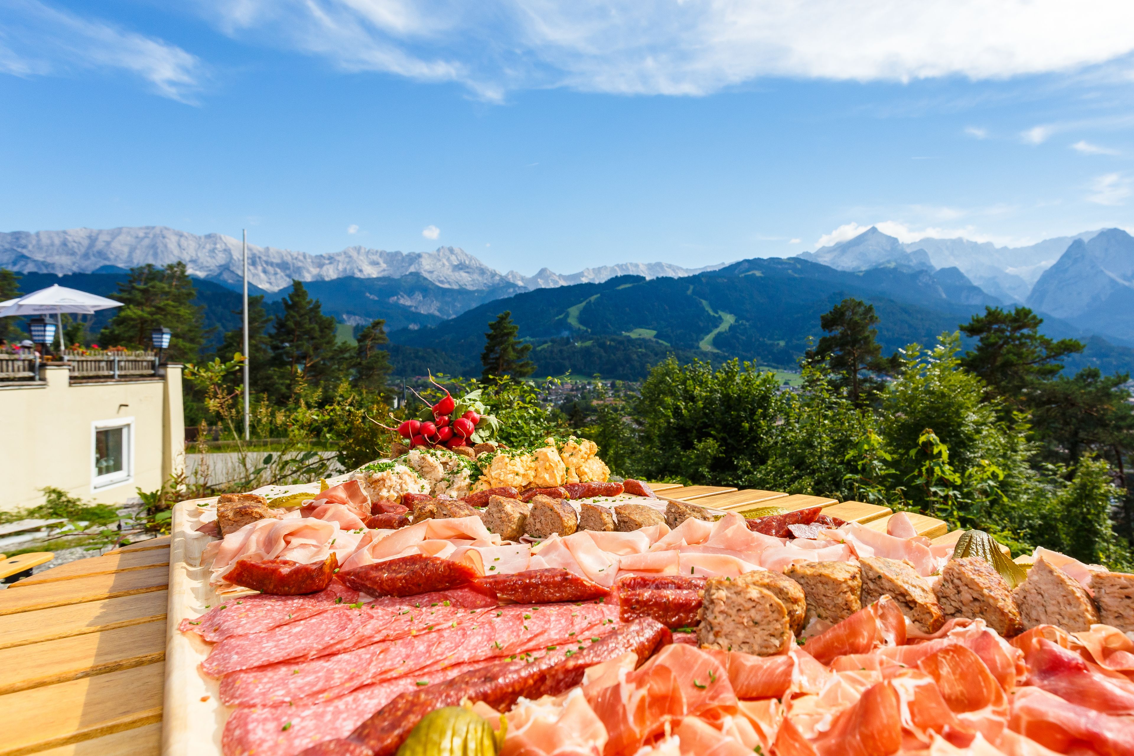 A charcuterie board with various meats and cheeses, set against a backdrop of mountains and a clear sky.