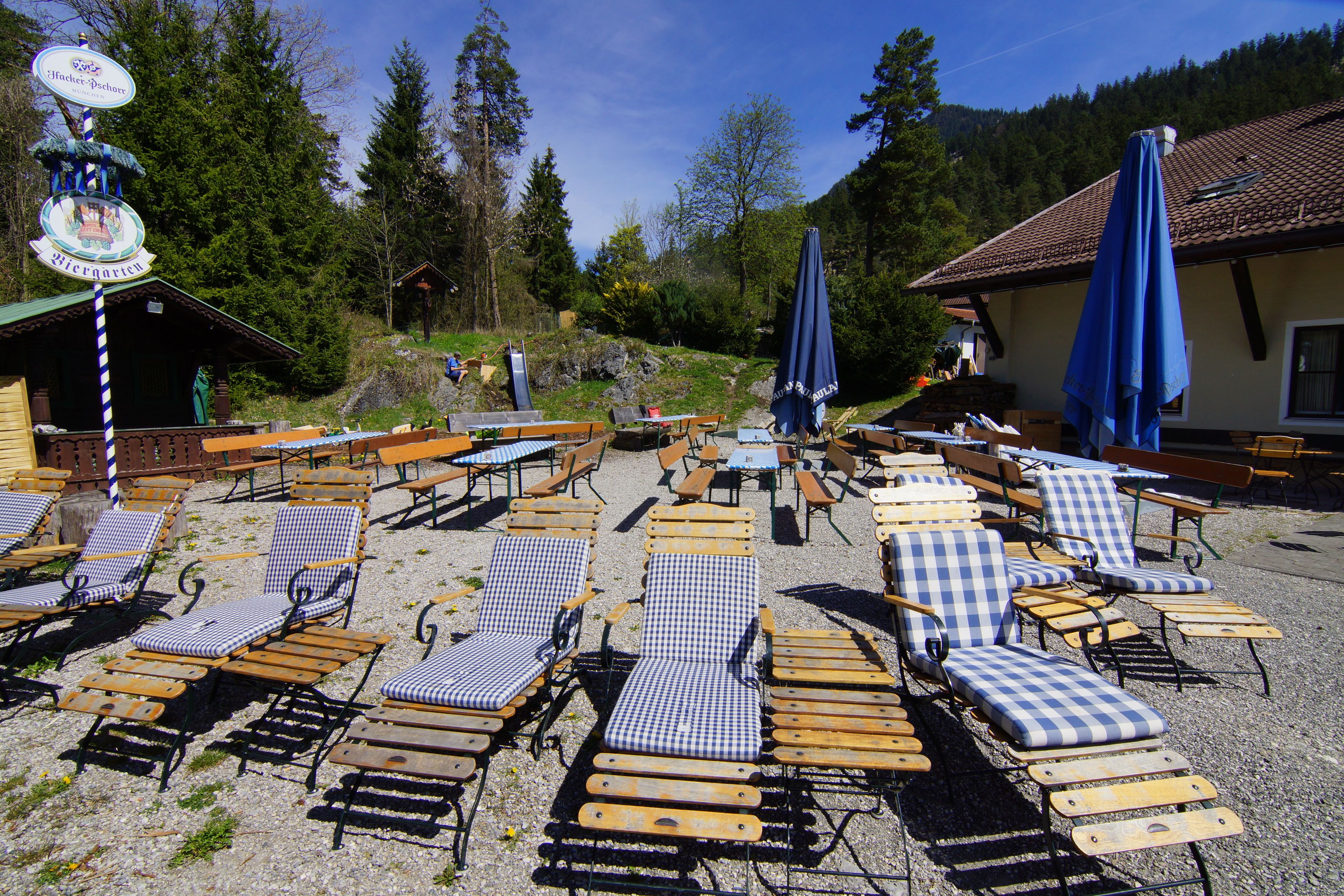 A sunlit outdoor seating area with checkered lounge chairs and tables, surrounded by trees and a building with a tiled roof.
