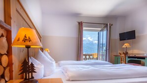 Une chambre d’hôtel avec un lit, une table de chevet en bois avec une lampe, un téléviseur fixé au mur et une vue sur les montagnes par la fenêtre.