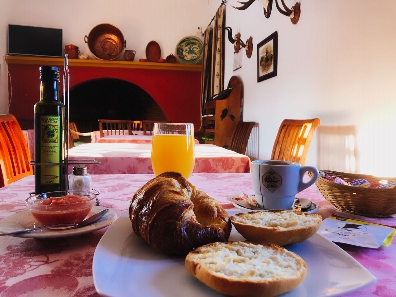 A breakfast setting with croissants, bread, a bottle of olive oil, a glass of orange juice, and a cup of coffee on a table with a red and white checkered tablecloth.