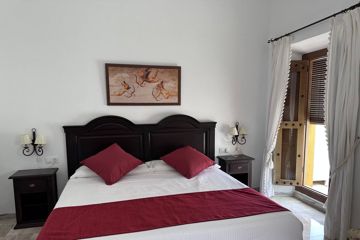 A neatly made bed with a red blanket and pillows, a dark wooden headboard, a wall-mounted painting, and a nightstand with a lamp.
