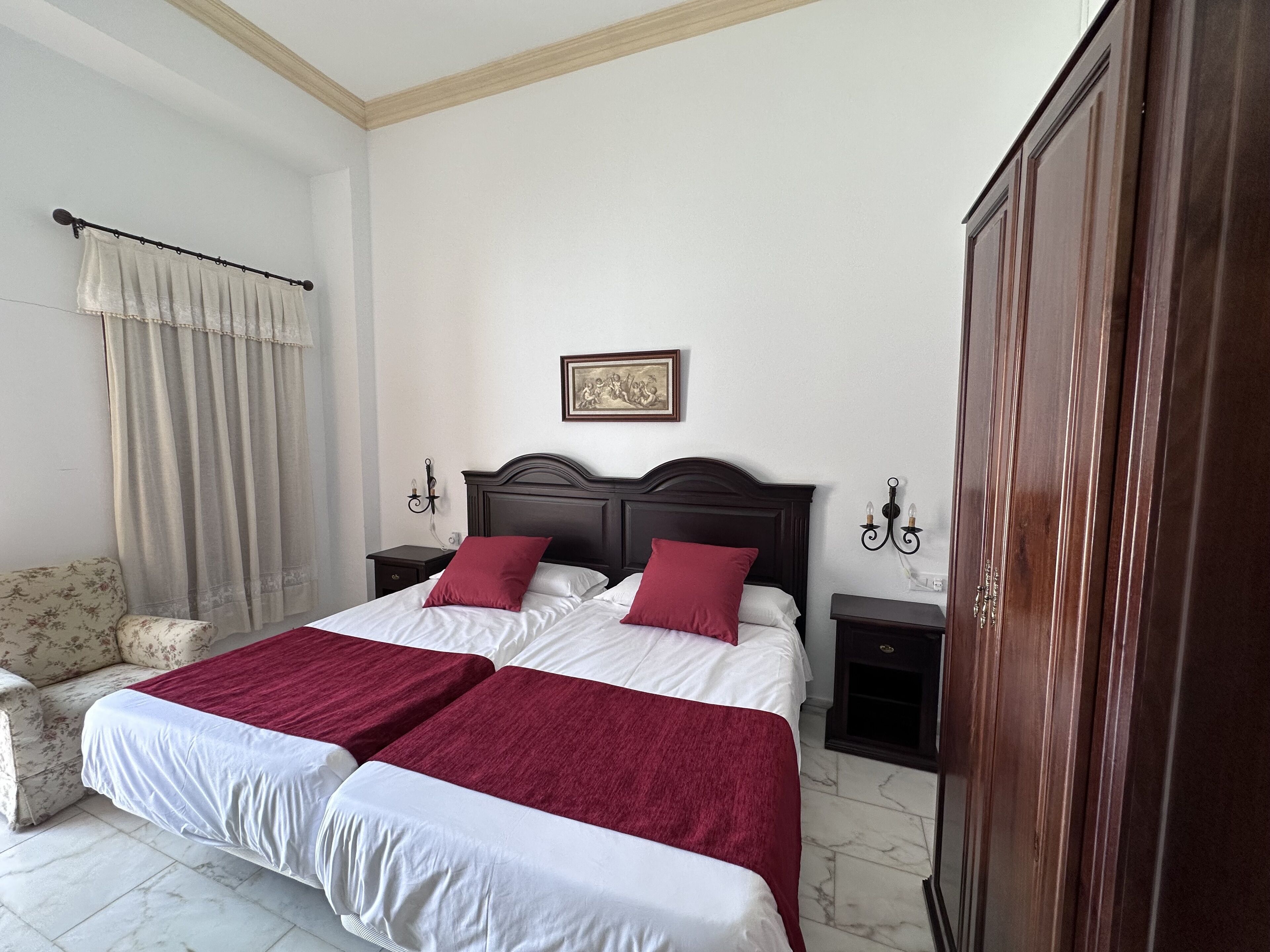 Una cama doble con ropa de cama blanca y roja, dos mesitas de noche con lámparas, un sillón con estampado floral, un armario de madera y un cuadro en la pared.