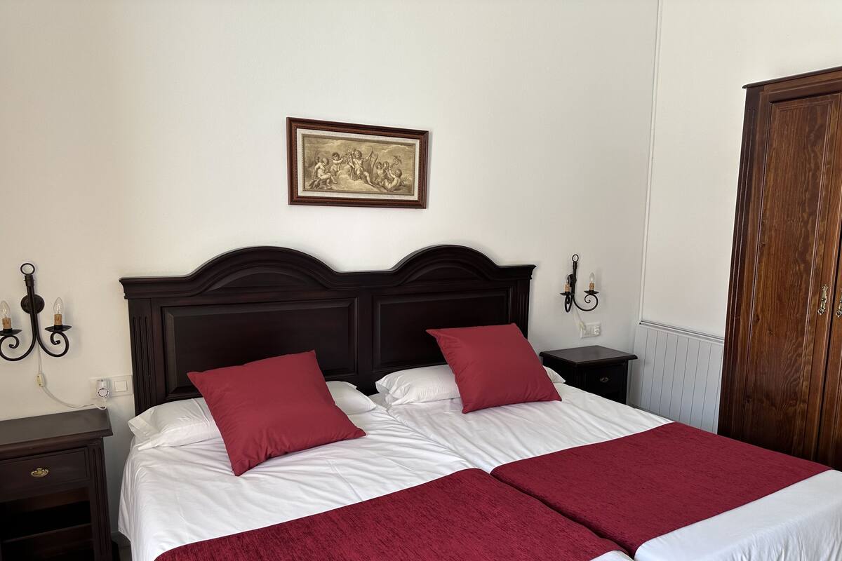 A double bed with white linens and red pillows, a dark wooden headboard, a wall-mounted picture, and a nightstand with a lamp.