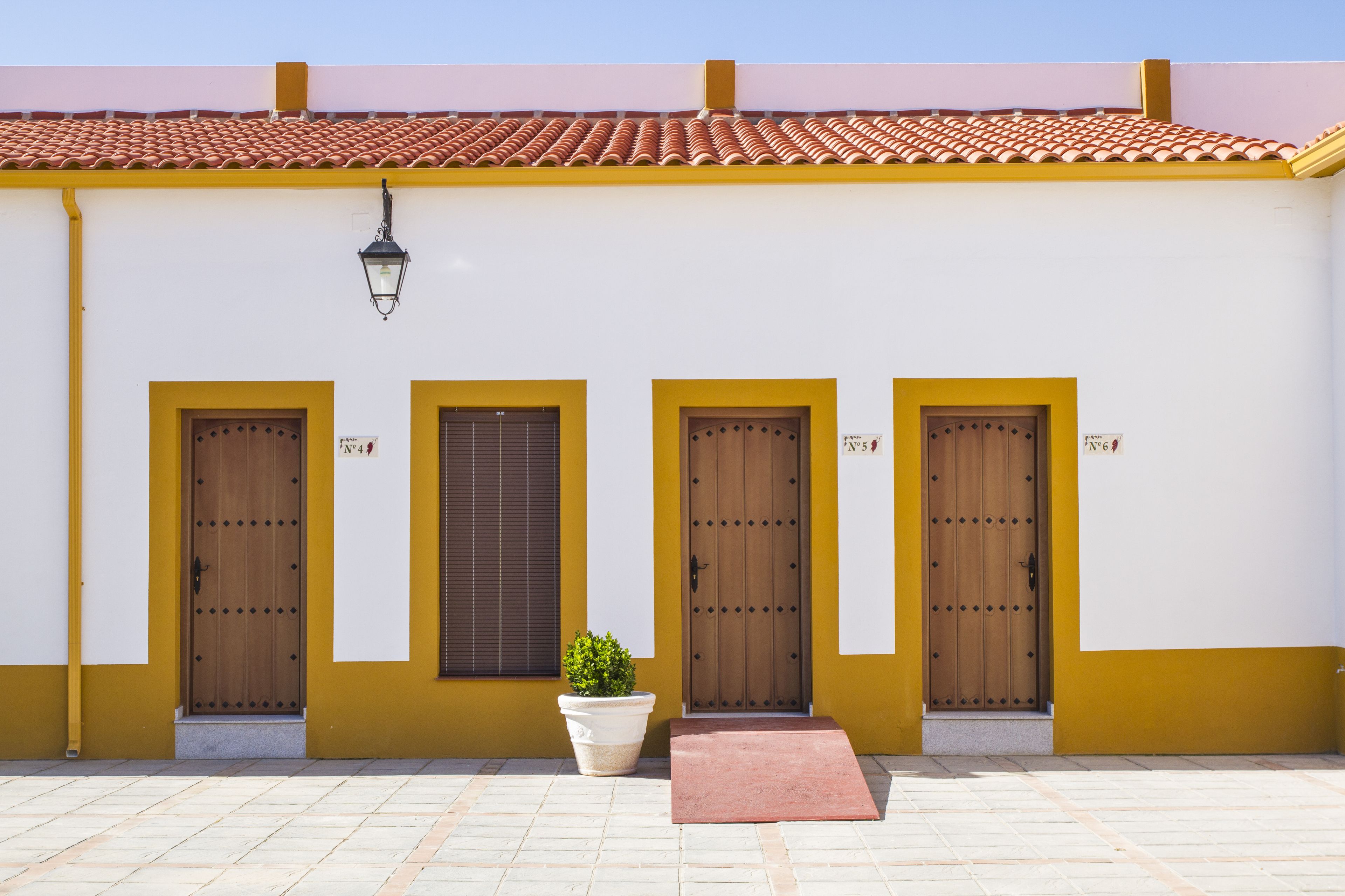 Un edificio con cuatro puertas, cada una marcada con un número. Hay una planta en maceta y un felpudo rojo frente a las puertas.