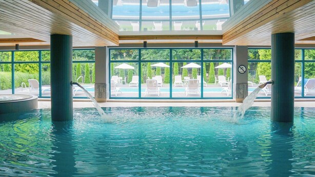 An indoor swimming pool with clear water, surrounded by large windows and white lounge chairs. - Hotel Villa Park Med & Spa (Ciechocinek)