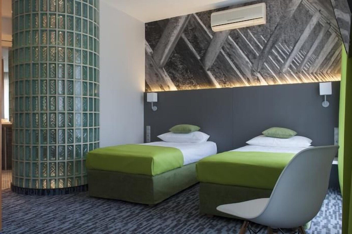 Habitación de hotel con dos camas, una pared de acento verde, alfombra estampada y un equipo moderno de aire acondicionado.