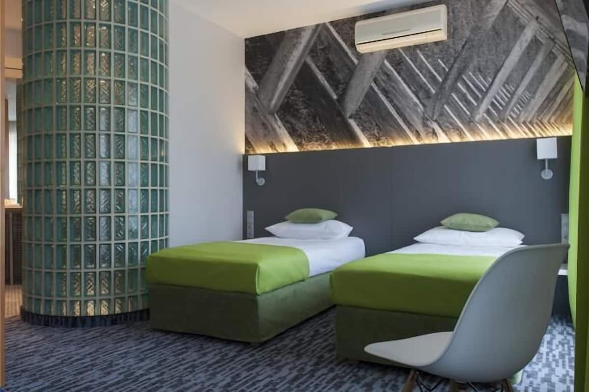 A hotel room with two beds, a green accent wall, a patterned carpet, and a modern air conditioning unit.