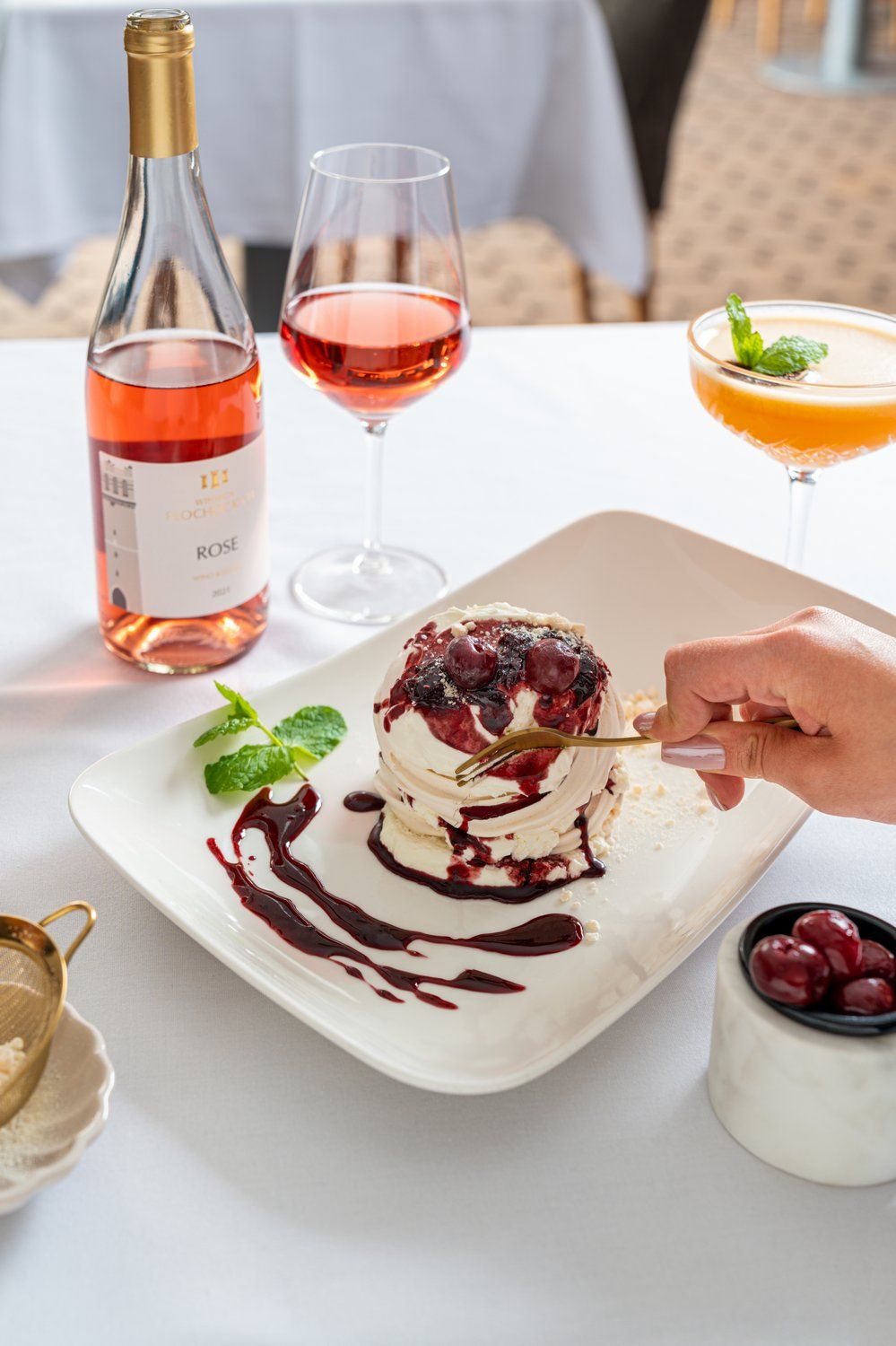 Un plato de postre con una bola de helado cubierto de bayas y un chorrito de salsa, una botella de vino rosado y una copa de vino rosado sobre una mesa.