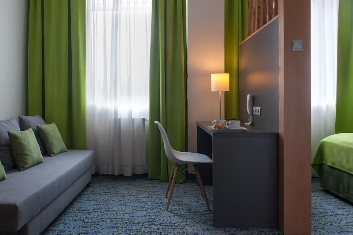 A hotel room with a grey sofa, a desk with a lamp, a chair, and a bed with green curtains.