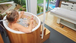 A spa tub filled with water and rose petals, a wooden floor, and a view of a pool and garden through large windows. - Hotel Villa Park Med & Spa (Ciechocinek)