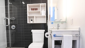 Un baño moderno con cabina de ducha de vidrio, inodoro blanco, lavabo con espejo y estante con artículos de aseo.