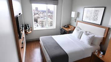 A hotel room with a bed, a desk, a chair, a TV, a window with a city view, and a framed artwork on the wall.