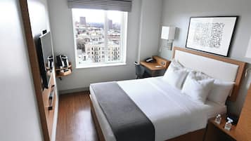 A hotel room with a bed, a desk, a chair, a TV, a bedside table, a lamp, a window with a city view, and a framed artwork on the wall.