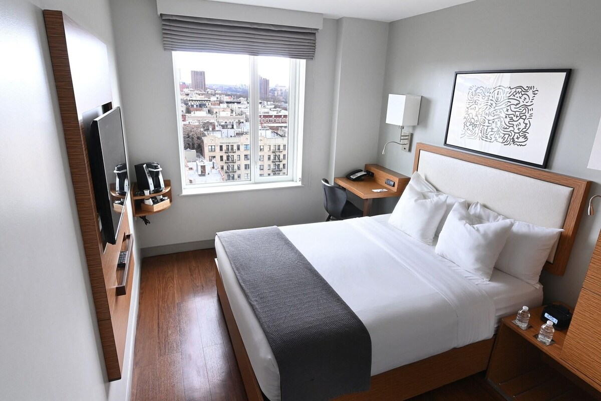Habitación de hotel con cama, escritorio, silla, televisión, ventana con vista a la ciudad y un cuadro en la pared.