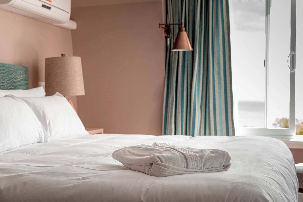 A neatly made bed with white linens and a folded towel, a lamp, and a window with curtains.
