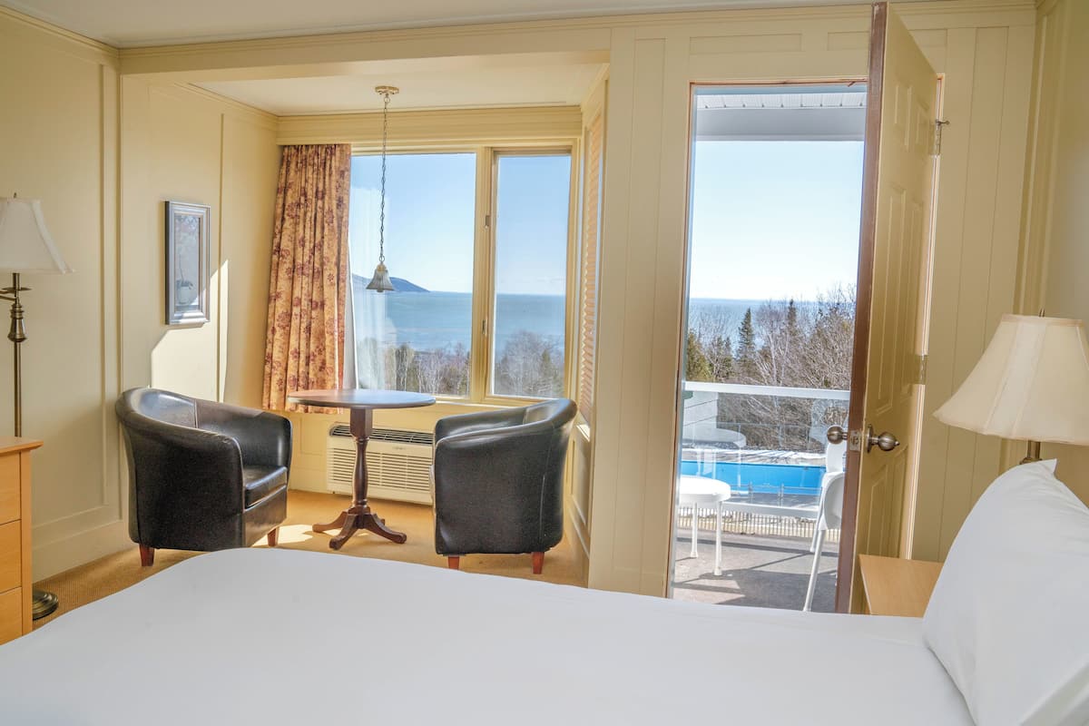 A hotel room with a bed, two chairs, a table, a lamp, a view of the sea, and a door leading to an outdoor area.