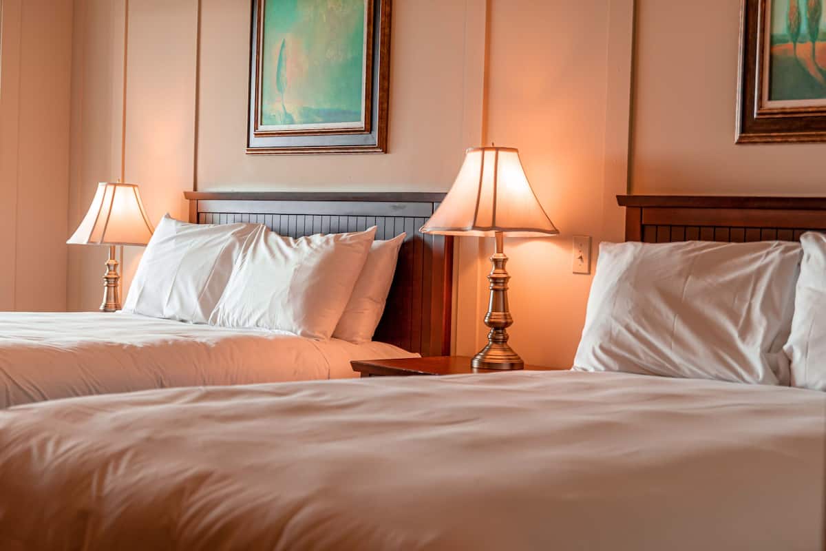 A hotel room with two beds, each with a wooden headboard, white bedding, and two bedside lamps.