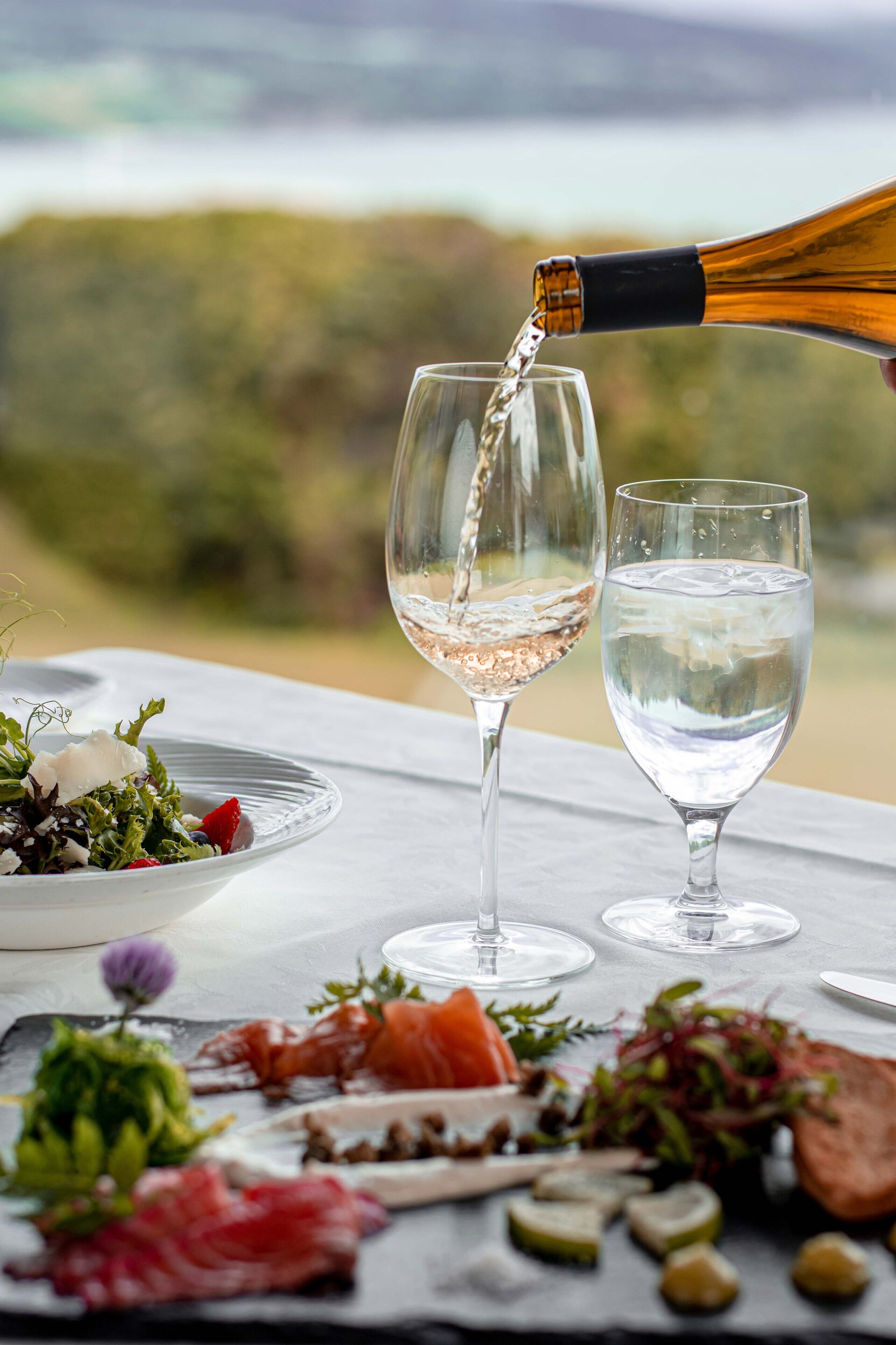 A table with a glass of white wine, a glass of water, and a variety of food including smoked salmon and vegetables.