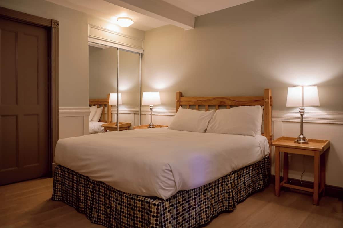 A neatly made bed with a wooden headboard, two bedside tables with lamps, a large mirror, and a wooden door.