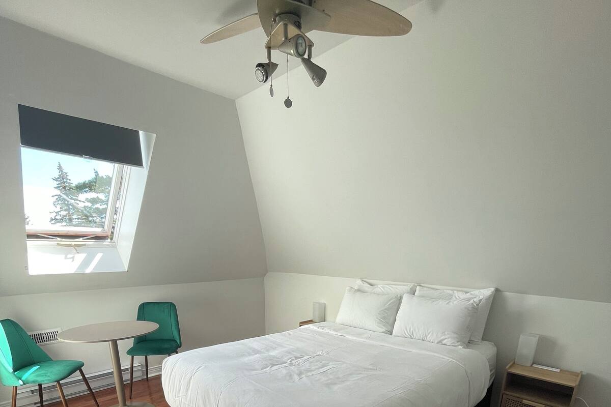 A bedroom with a bed, a desk, two chairs, a ceiling fan, a nightstand, and a window with a view of trees.