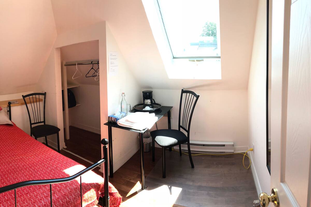A bedroom with a bed, a desk, and a chair. There is a window providing natural light and a view of the outside.