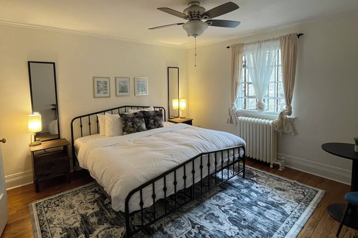 A bedroom with a bed, a ceiling fan, a nightstand with a lamp, a window with curtains, and a patterned rug.