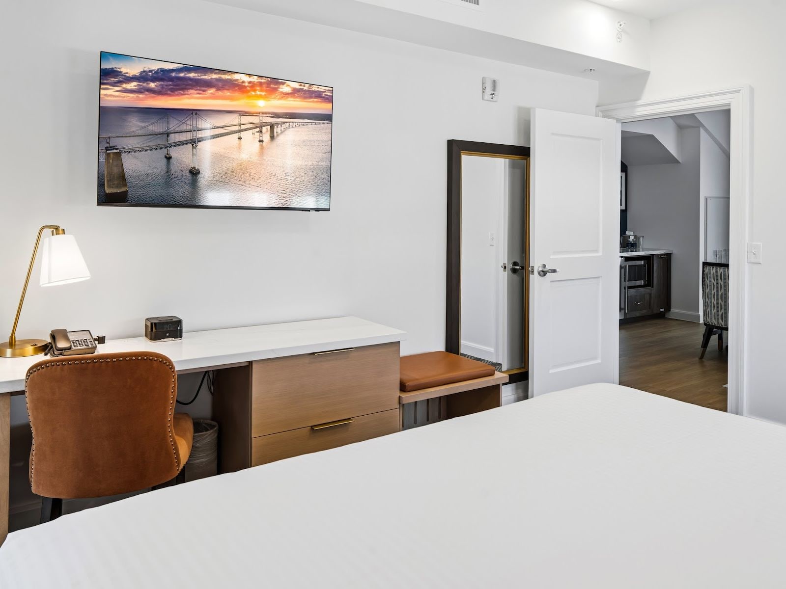 A hotel room with a bed, desk, chair, and a painting of a sunset over a bridge.