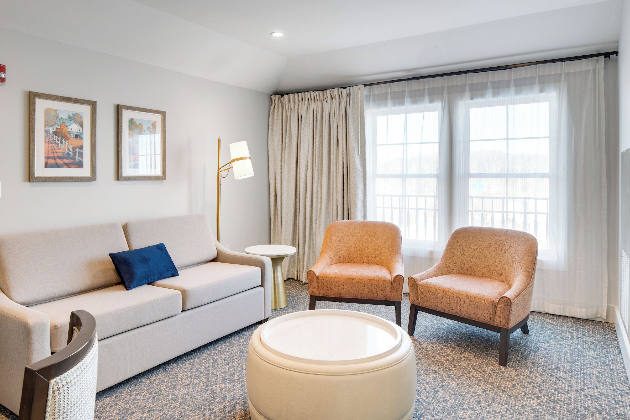 A hotel room with a sofa, two armchairs, a small round table, and a lamp. There are two framed pictures on the wall.