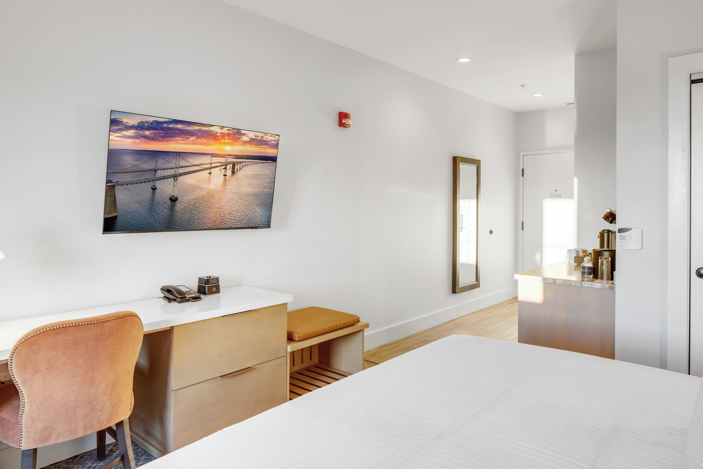 A modern hotel room with a large bed, a desk with a chair, a small bench, a painting of a sunset over a bridge, and a mirror on the wall.