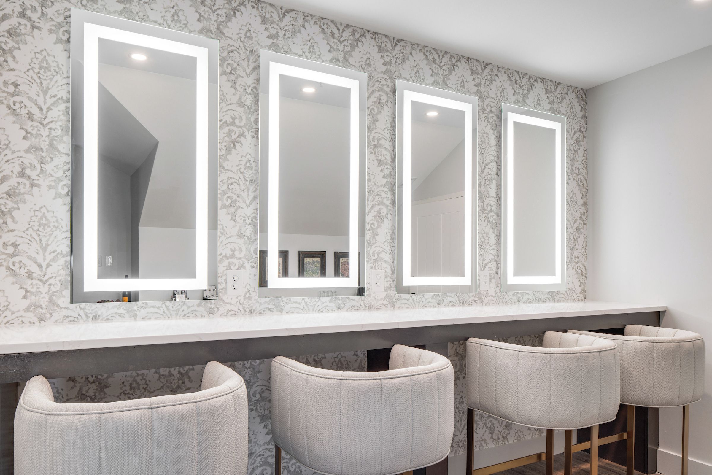 Une salle de bain avec trois miroirs fixés au mur, un meuble-lavabo doté d’un plan en marbre et quatre tabourets de bar rembourrés.